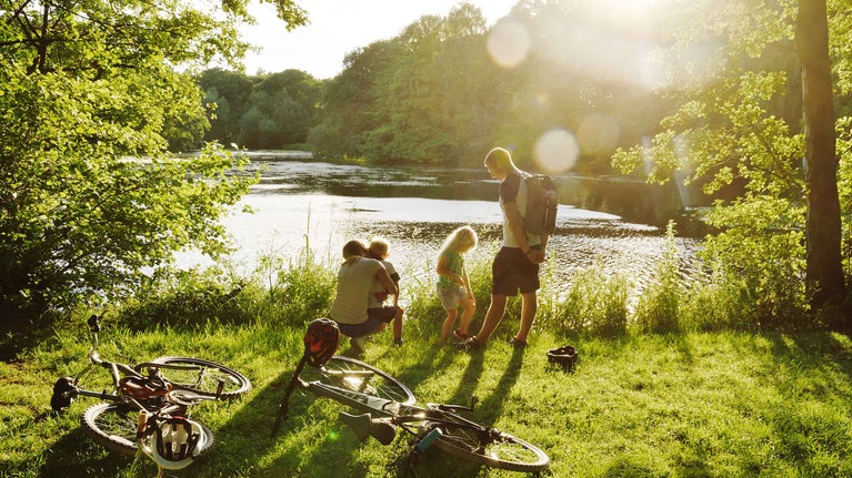 A family stand by the water's edge in the parkland at Nostell Priory, with their bikes on the floor next to them